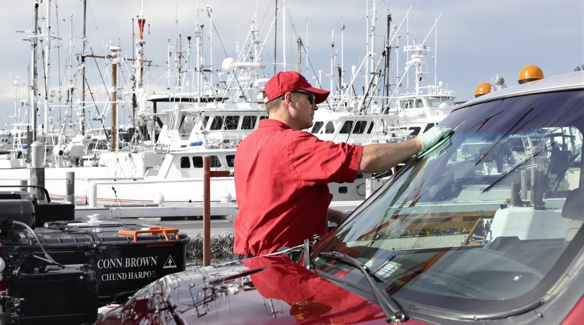Auto glass technician in red uniform working on windshield of vehicle at Conn Brown Harbor Aransas Pass Texas with fishing boats in background