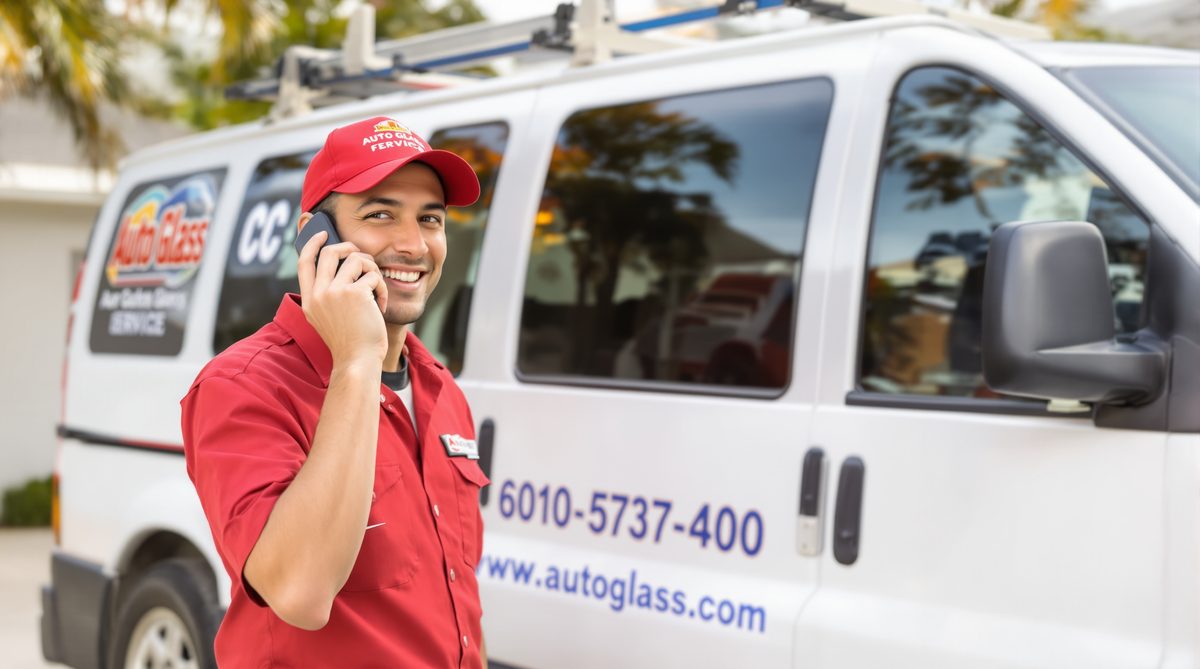 Friendly CC Auto Glass Pros technician in red uniform talking on phone while standing in front of white service van with CC Auto Glass branding